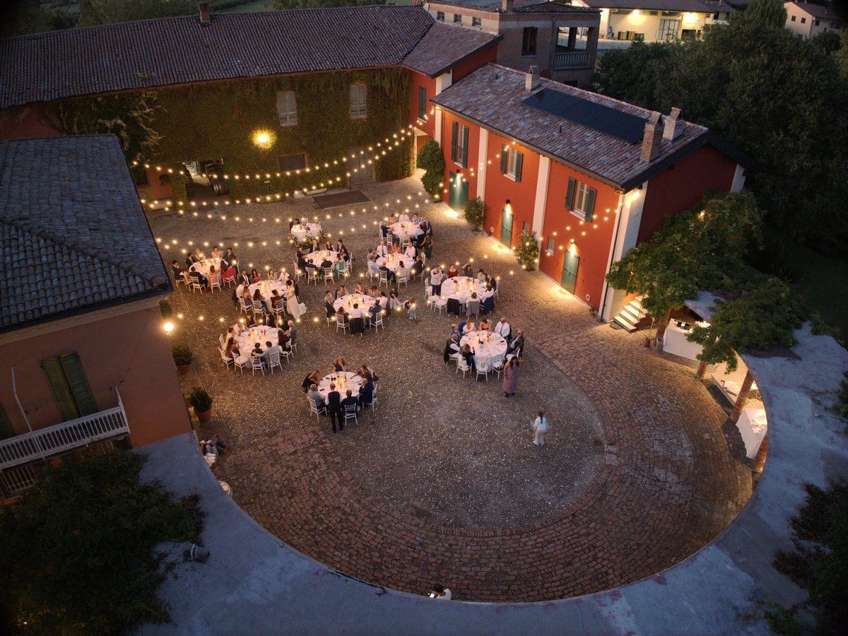 La tenuta vista dal drone al tramonto: la casa rossa, la corte in acciottolato con le file di luci e gli invitati a cena ai tavoli rotondi